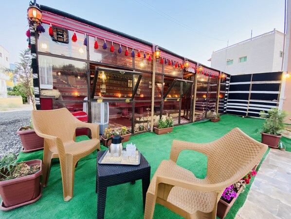 An outdoor seating area with wicker chairs and a table, surrounded by potted plants and a green carpet.