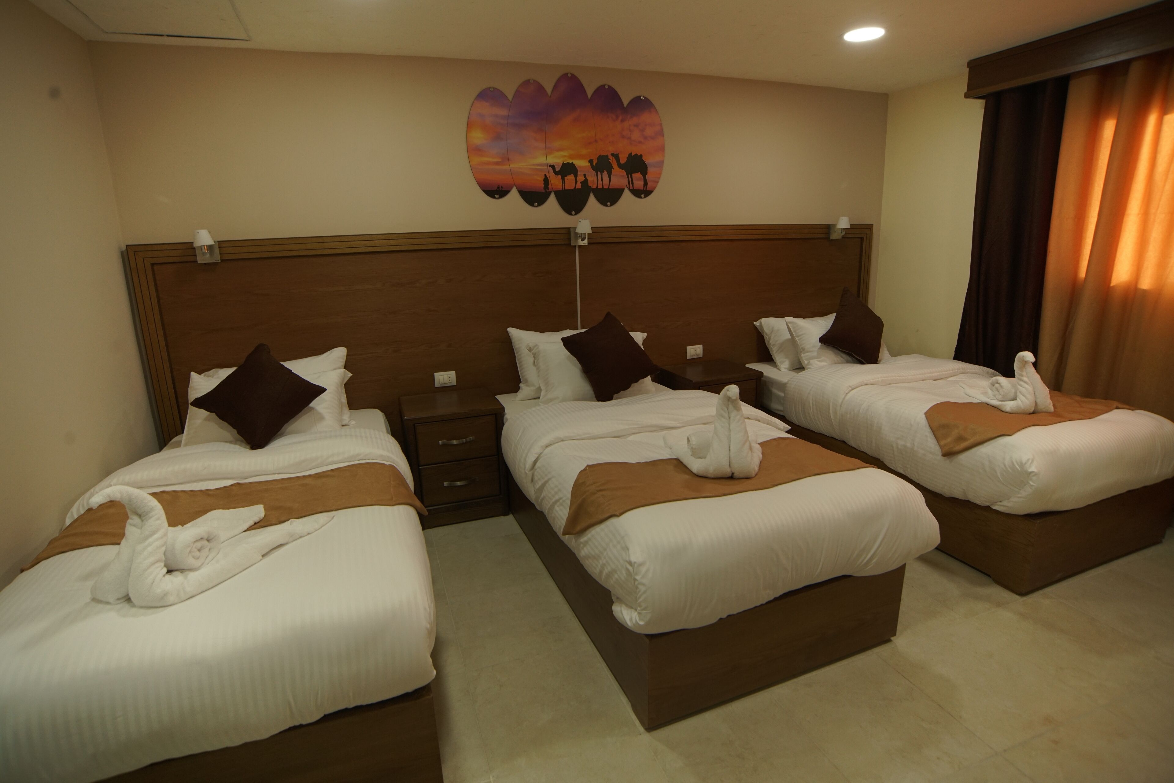 A hotel room with two beds, each with a wooden headboard, white bedding, and brown throw blankets. There is a wall art depicting a desert scene with camels.