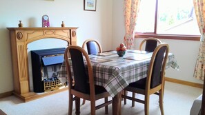 A dining area with a fireplace, wooden chairs, a plaid tablecloth, and a window with floral curtains. - 2 Bed Home With Private Garden in the Highlands (Beauly)