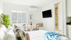 A bedroom with a bed, a desk, chairs, a TV, and a window with a view of buildings. - Kasa Del Ray Alexandria (Alexandria)