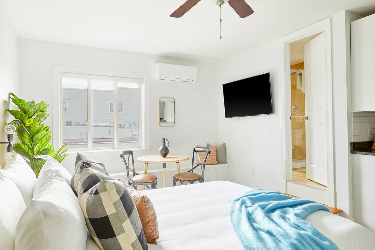 Une chambre à coucher comprenant un lit, un bureau, des chaises, un téléviseur et une fenêtre donnant sur des immeubles.