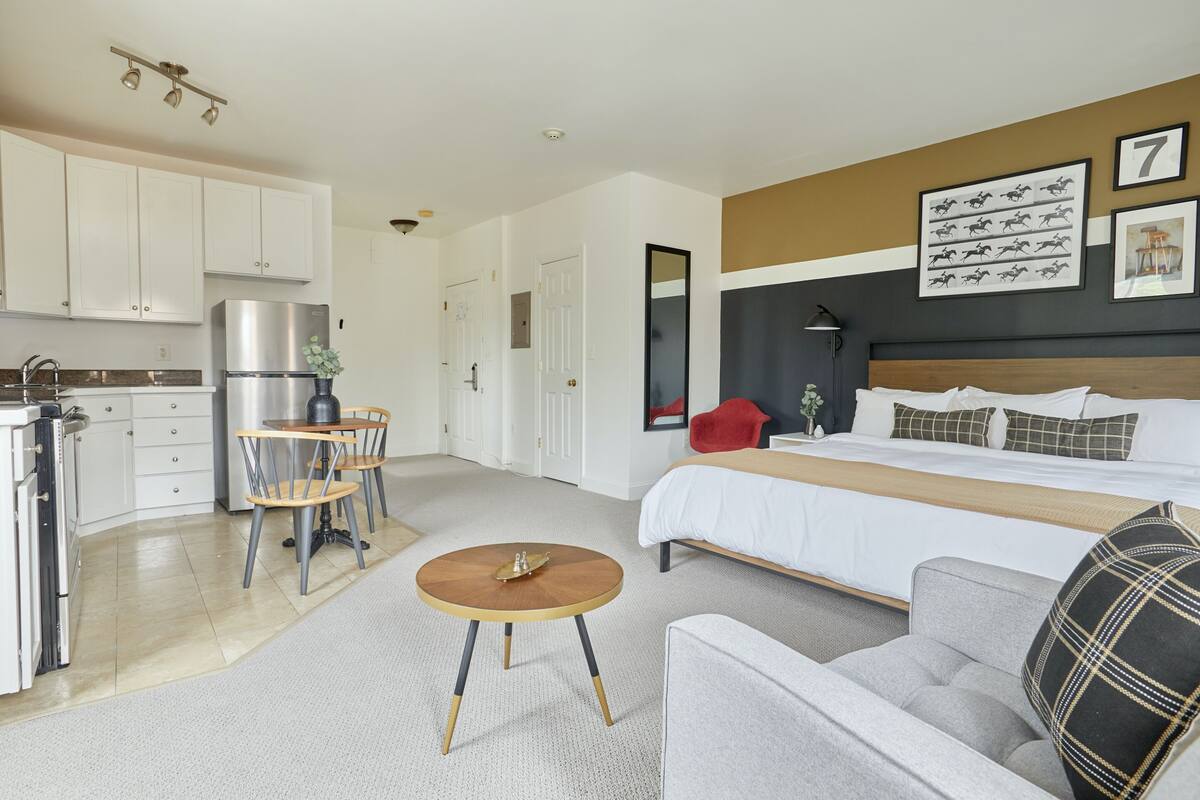 Une chambre d’hôtel moderne comprenant un lit, un canapé, un coin repas avec des chaises, une cuisine avec un réfrigérateur et un couloir.