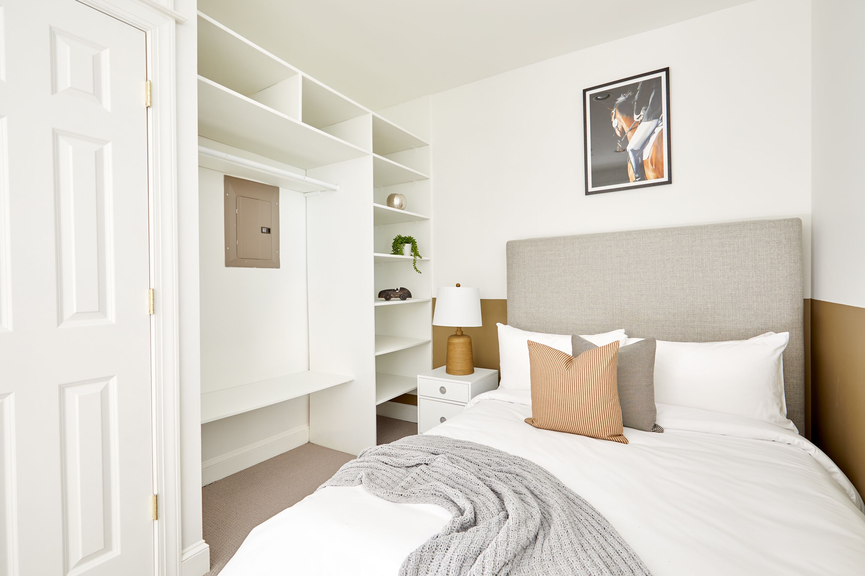 Un dormitorio con cama, mesita de noche, lámpara, un cuadro en la pared y un armario con estantes.