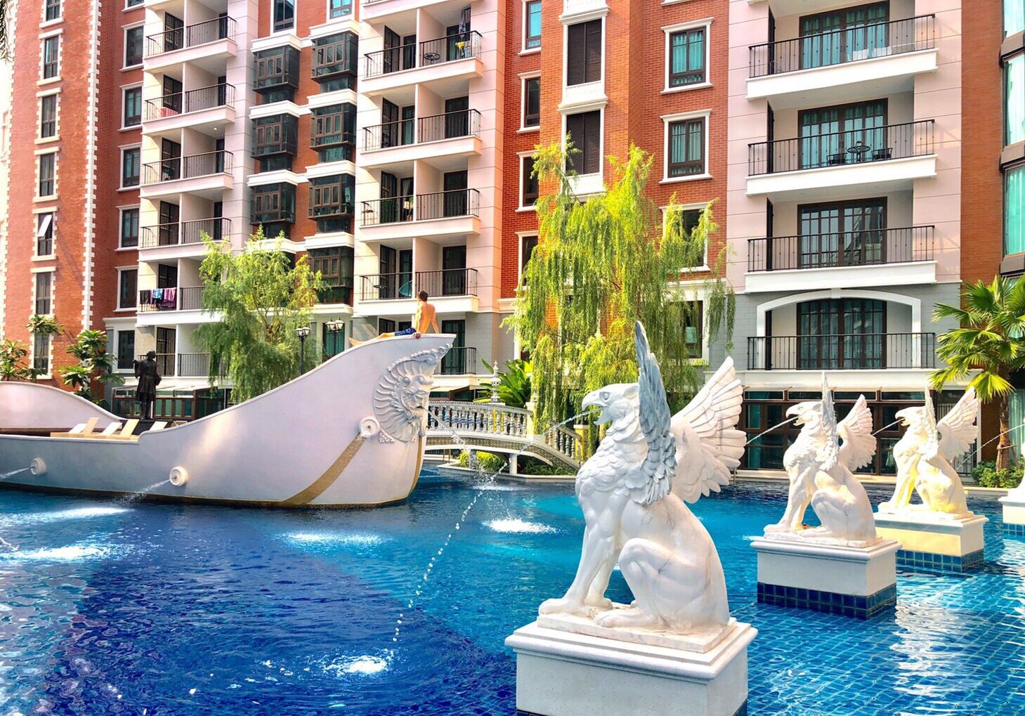 A hotel pool area with a large decorative ship, statues, and a multi-story building with balconies in the background.