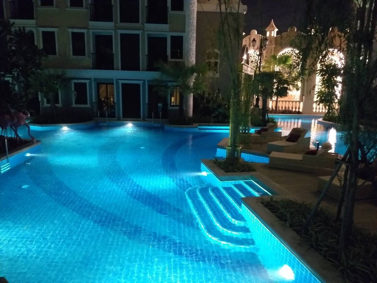 Una piscina nocturna con bordes iluminados, rodeada de sillones de descanso y un edificio con varias ventanas.