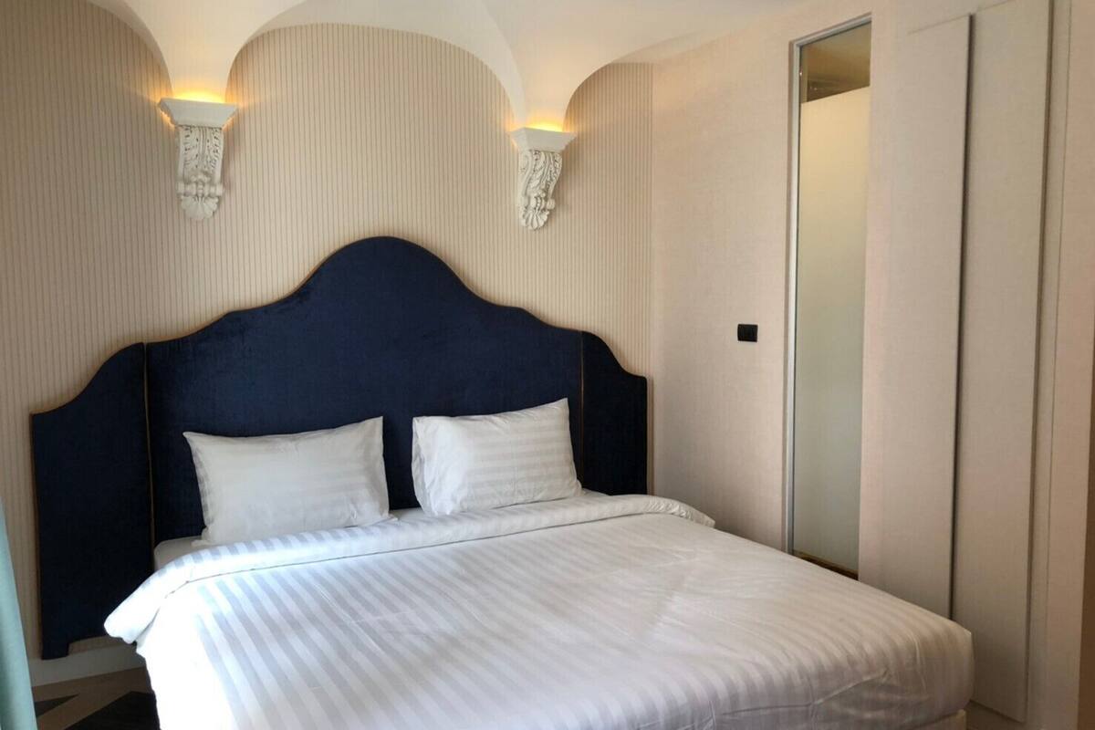 A neatly made bed with a dark headboard and two pillows, a light-colored striped bedspread, and a wall-mounted light fixture.