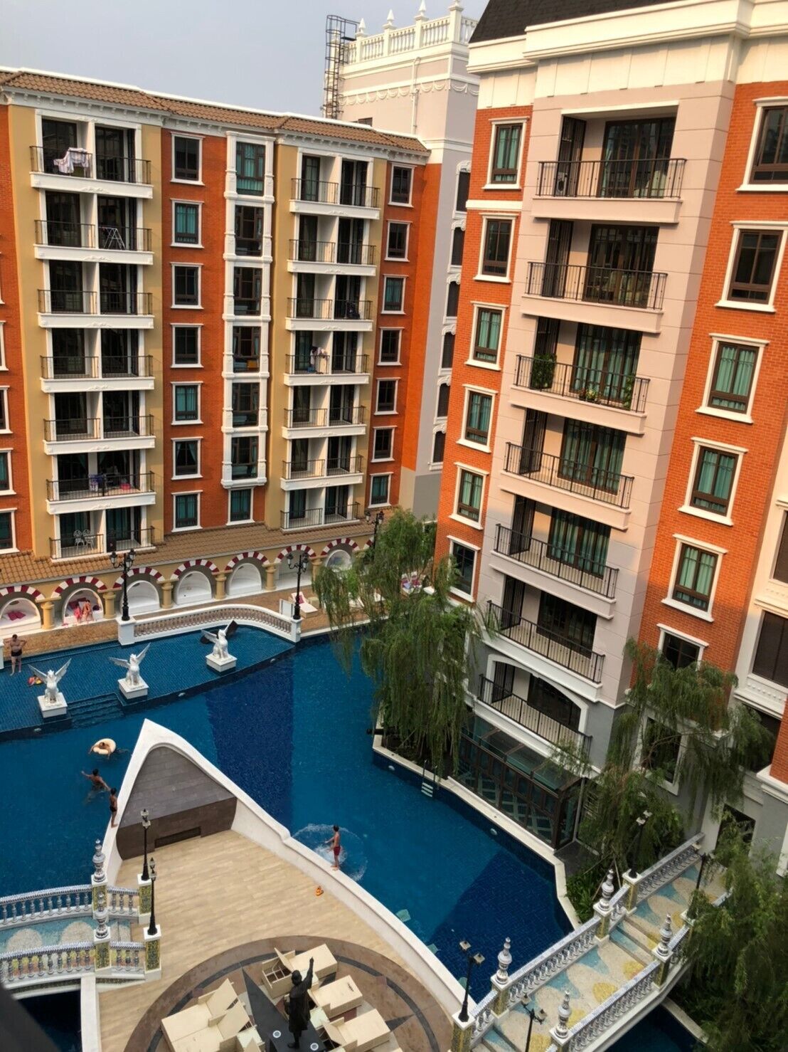 A multi-story residential building with balconies, a swimming pool, and a landscaped area with statues.