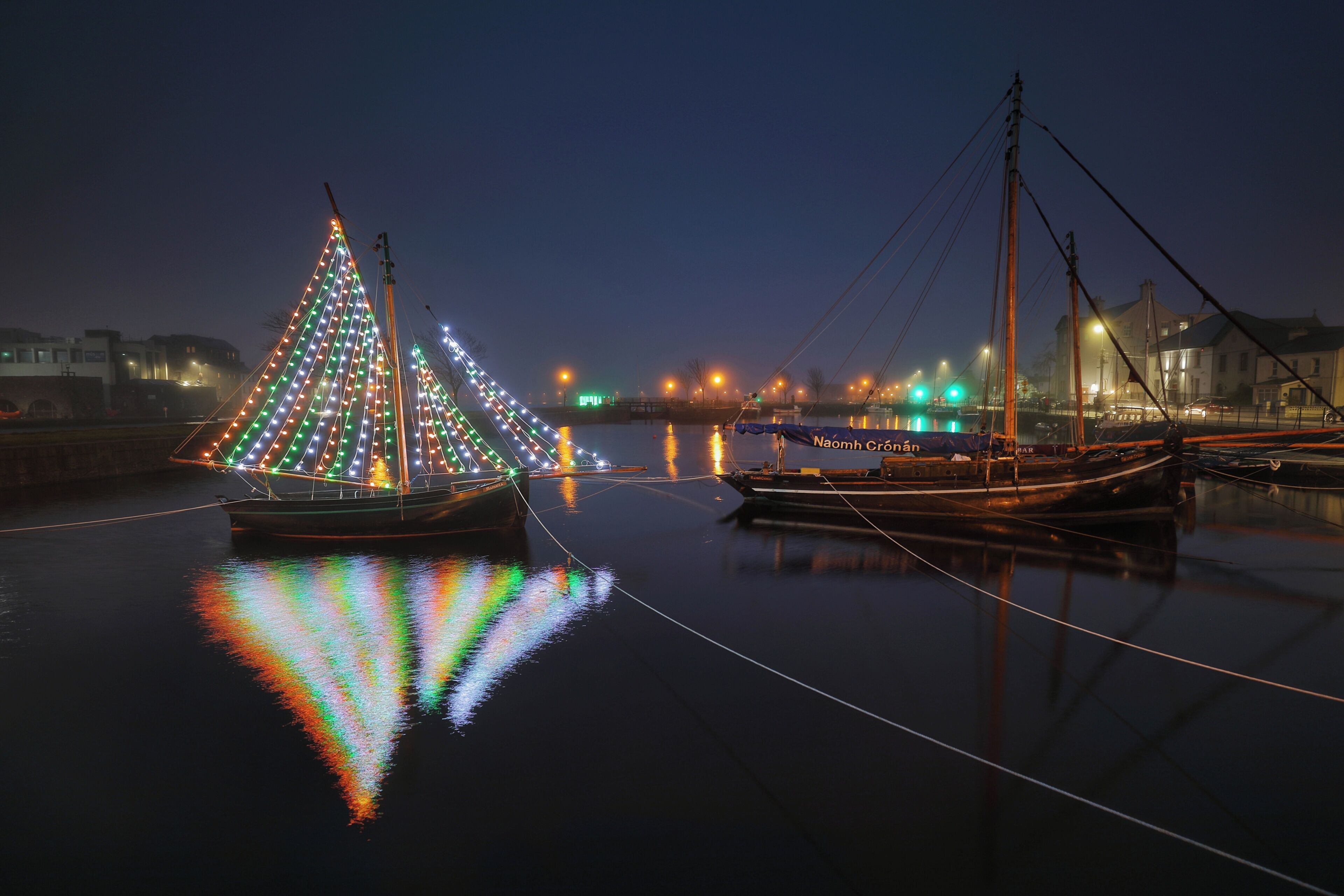 Port a la nit, amb embarcacions il·luminades i una superfície d'aigua tranquil·la que reflecteix llums.