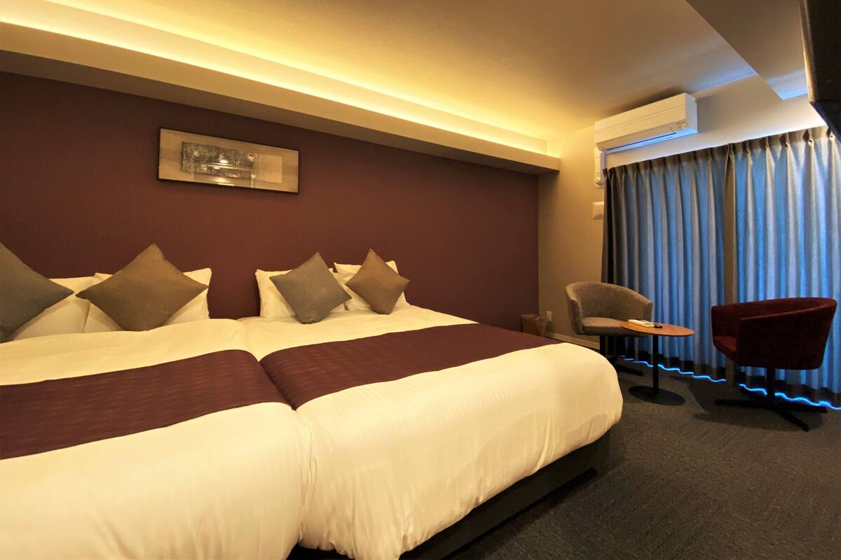 A hotel room with a large bed, a chair, an air conditioning unit, and a window with curtains.