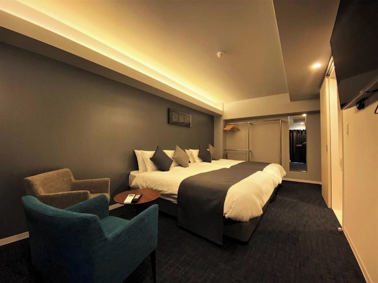 Quarto de hotel com cama, uma cadeira, uma mesinha e um guarda-roupa.
