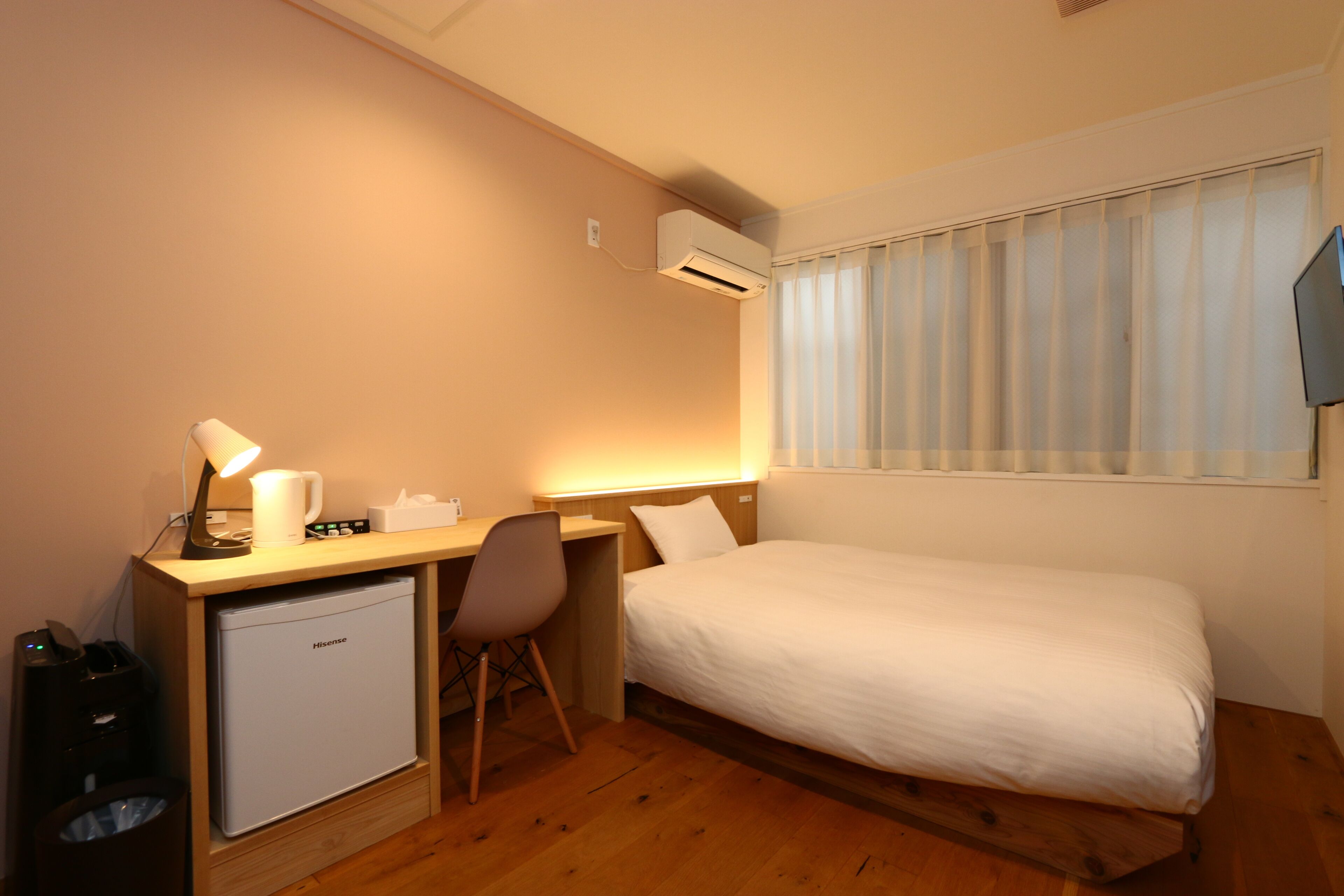 A compact hotel room with a single bed, a small desk with a chair, a desk lamp, a mini fridge, and a window with blinds.