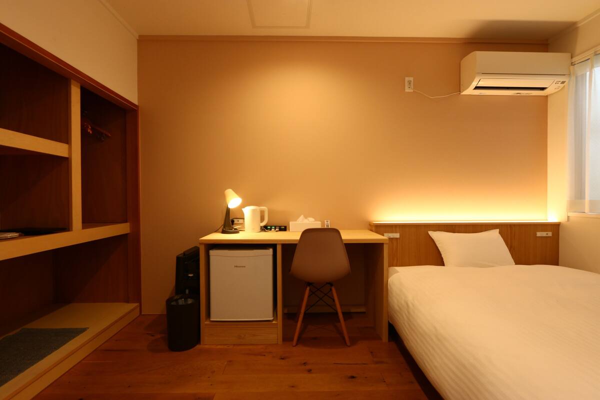 A compact hotel room with a single bed, a desk with a chair, a small refrigerator, and an air conditioning unit mounted on the wall.