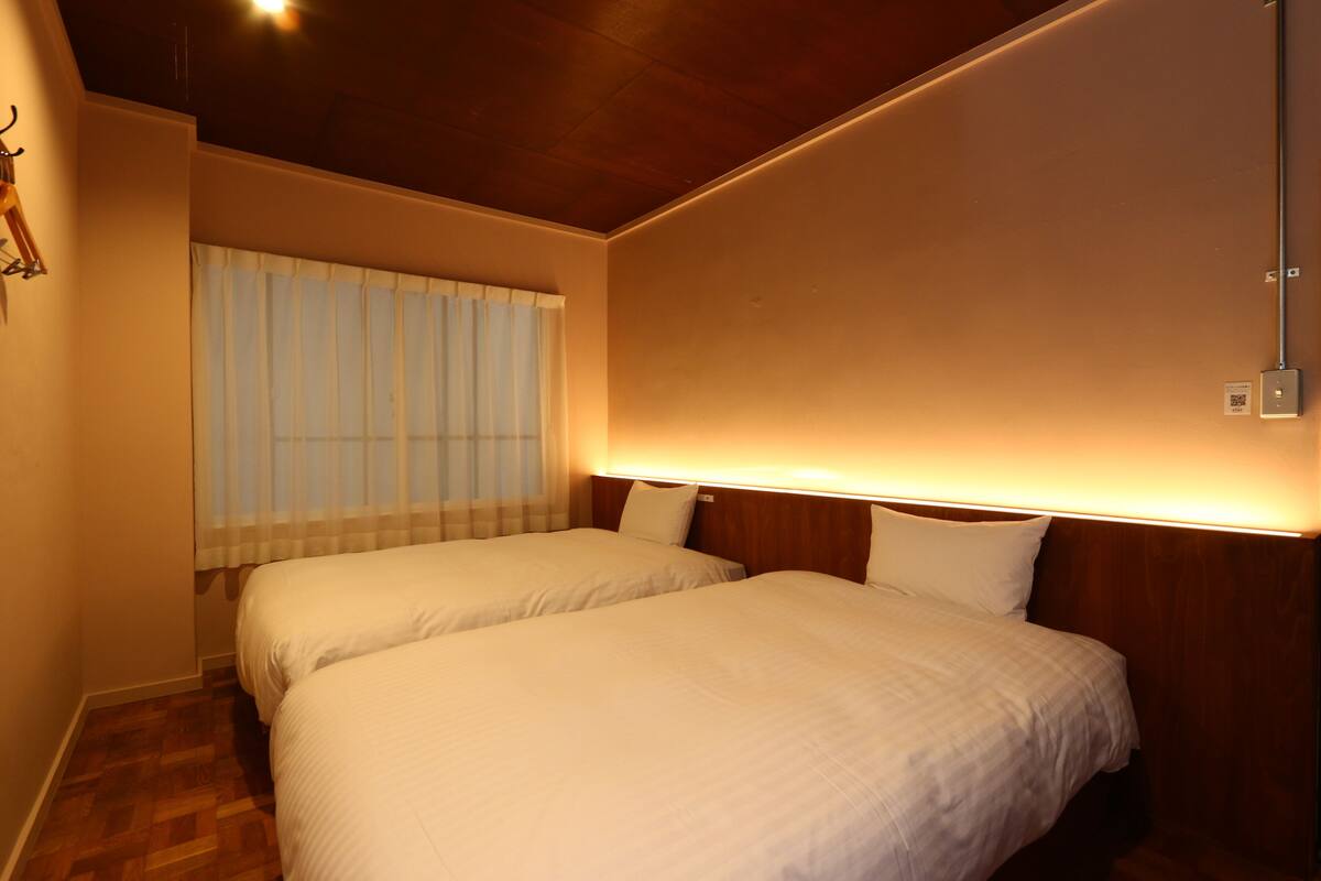 A hotel room with two beds, wooden headboards, a window with sheer curtains, and a wall-mounted light fixture.