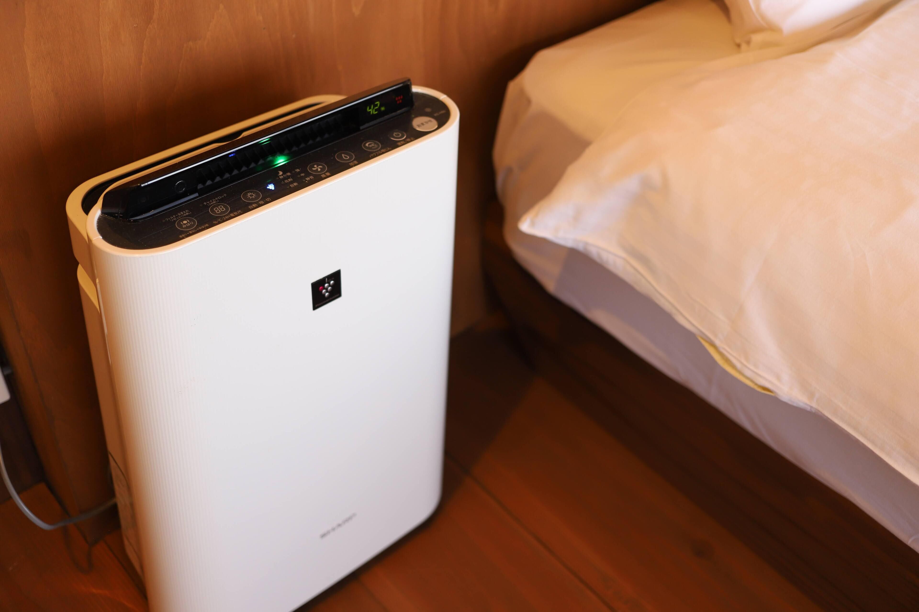 Un aire acondicionado portátil con pantalla digital, ubicado cerca de una cama en una habitación con piso de madera.