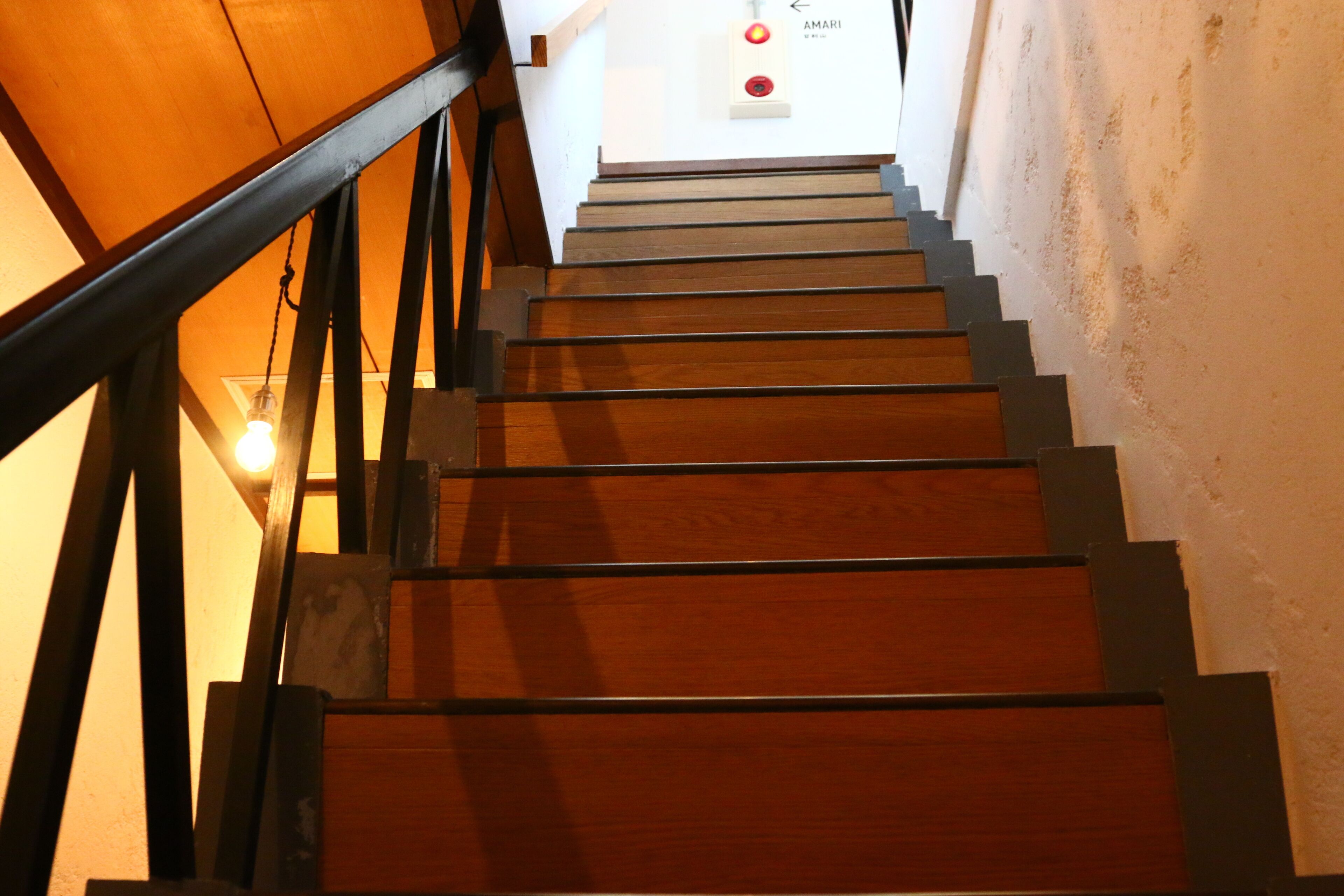 A wooden staircase with a metal handrail leading to a landing with a light switch and a wall-mounted light fixture.