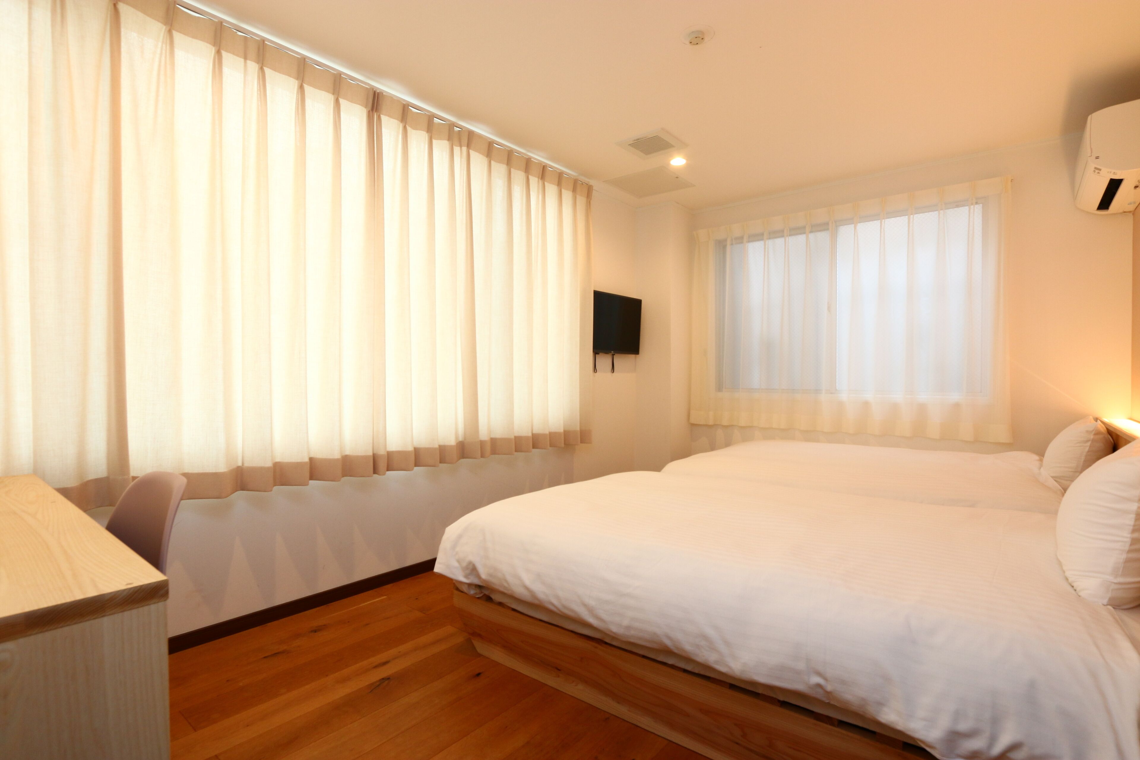 A bedroom with a large bed, wooden flooring, a desk, and a TV mounted on the wall.