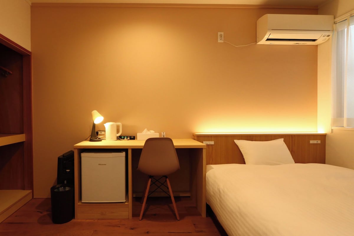 Una habitación de hotel con una cama, un escritorio con lámpara, un calefactor pequeño y un aire acondicionado.