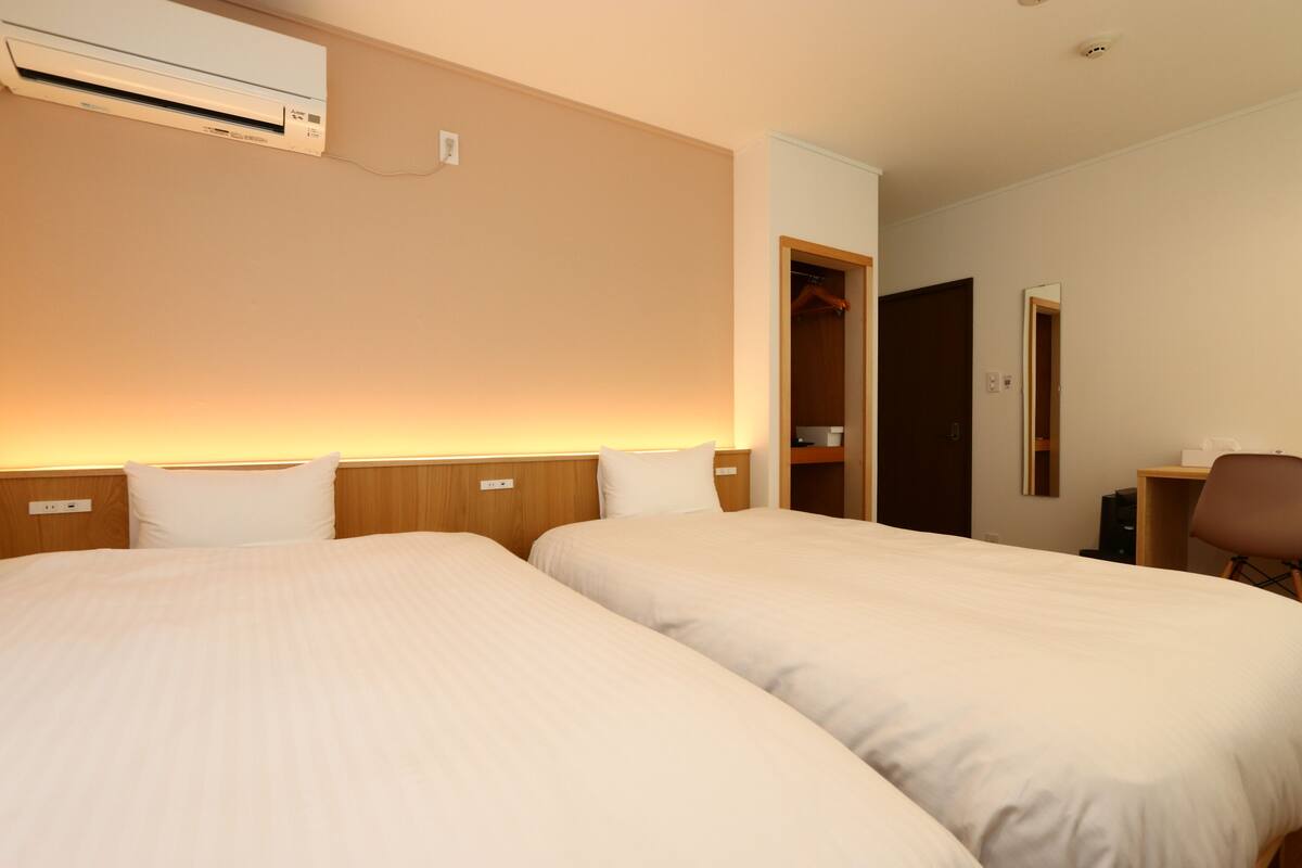A hotel room with two beds, a wardrobe, a desk, and an air conditioning unit.