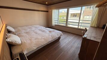 Ein Schlafzimmer mit einem großen Bett, Holzboden, einem Fenster mit Blick auf Gebäude und einem kleinen Tisch mit einem Gerät darauf.