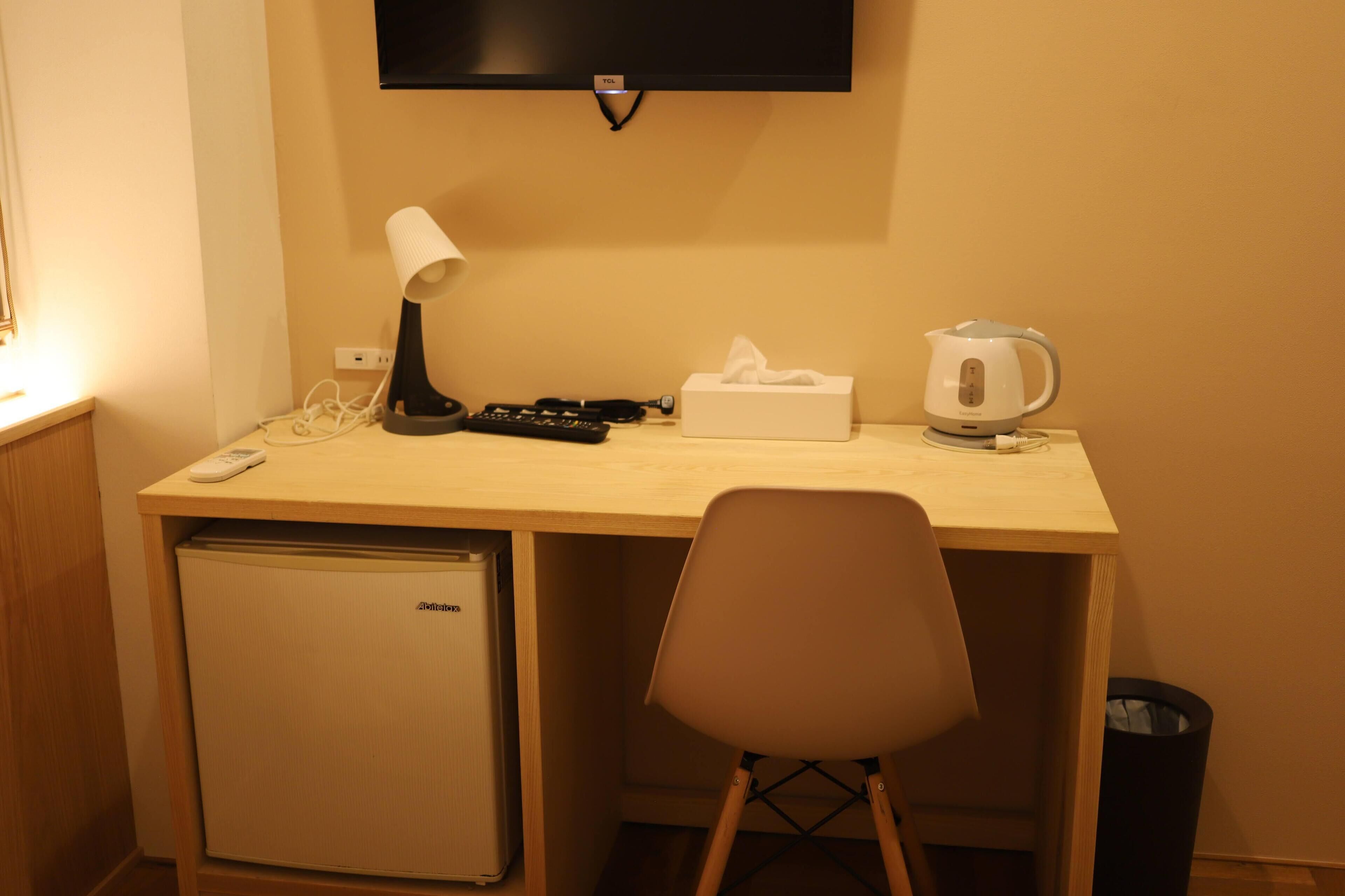A compact hotel room with a wooden desk, a small refrigerator, a chair, a television, a lamp, a remote control, a tissue box, and a kettle.