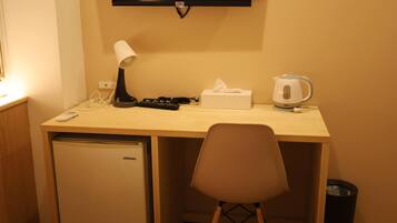 A compact hotel room with a wooden desk, a small refrigerator, a chair, a television, a lamp, a remote control, a tissue box, and a kettle.