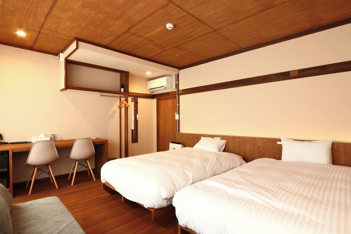 A hotel room with two beds, a desk, chairs, and an air conditioning unit.