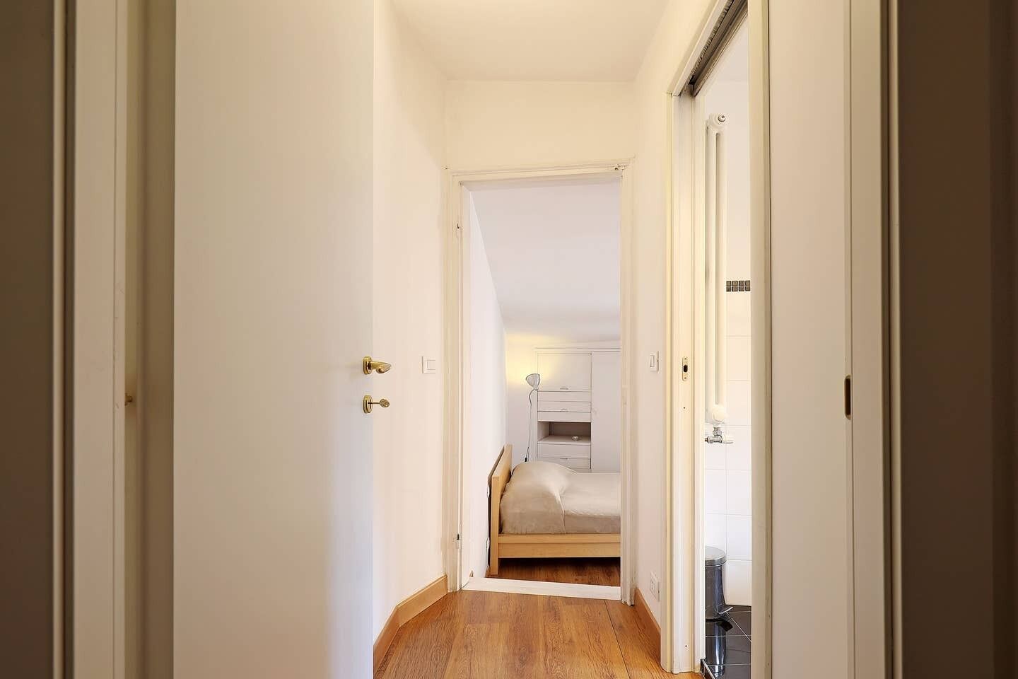 A narrow hallway leading to a bedroom with a bed, bedside table, and a lamp.
