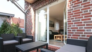 A patio with wicker furniture, a wooden table, and a glass door leading to an interior space with a sofa and a desk. - Vacation breathing space in Greetsiel (Greetsiel)