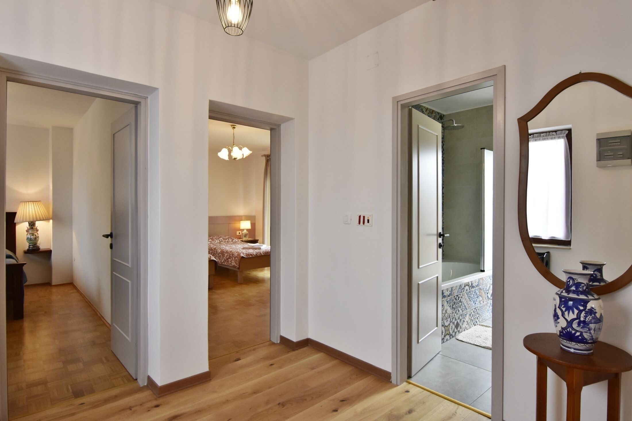 A hallway with wooden flooring leading to a bedroom with a bed and a nightstand. There is a mirror on the wall and a vase on a small table.