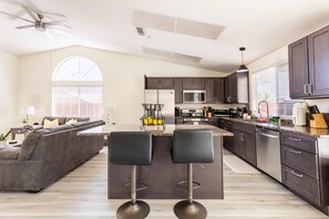 Une cuisine moderne avec des meubles de couleur sombre, un îlot central doté de tabourets, et un coin repas comprenant un canapé et un ventilateur de plafond.