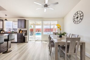 A modern kitchen with dark cabinets, a dining area with wooden chairs, a ceiling fan, and a wall clock. - Salt Water Pool & Spa with Mountain Sunset Views! (Desert Hot Springs)