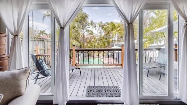 A room with a view of a wooden deck and a pool, curtains drawn to one side, and a chair visible in the foreground. - The Periwinkle House – Bask Poolside at this Sunlit West Palm Retreat (West Palm Beach)