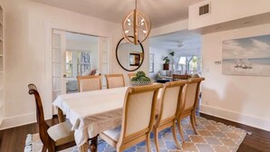 A dining area with a table set for four, wooden chairs, a chandelier, and a patterned rug. - The Periwinkle House – Bask Poolside at this Sunlit West Palm Retreat (West Palm Beach)