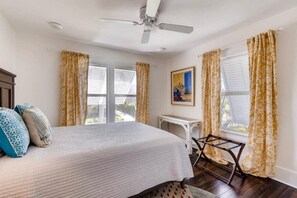A bedroom with a bed, curtains, a ceiling fan, a window, and a painting on the wall. - The Periwinkle House – Bask Poolside at this Sunlit West Palm Retreat (West Palm Beach)