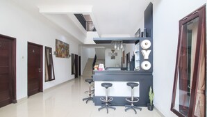 A modern hotel lobby with a reception desk, three bar stools, a staircase, and a large mirror.