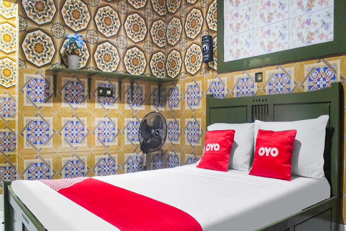 Cama com roupa de cama branca e vermelha, um ventilador e uma parede com azulejos estampados.