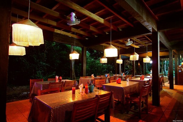 Un área de comedor al aire libre con mesas y sillas, cubierta por una estructura de madera con luces colgantes.
