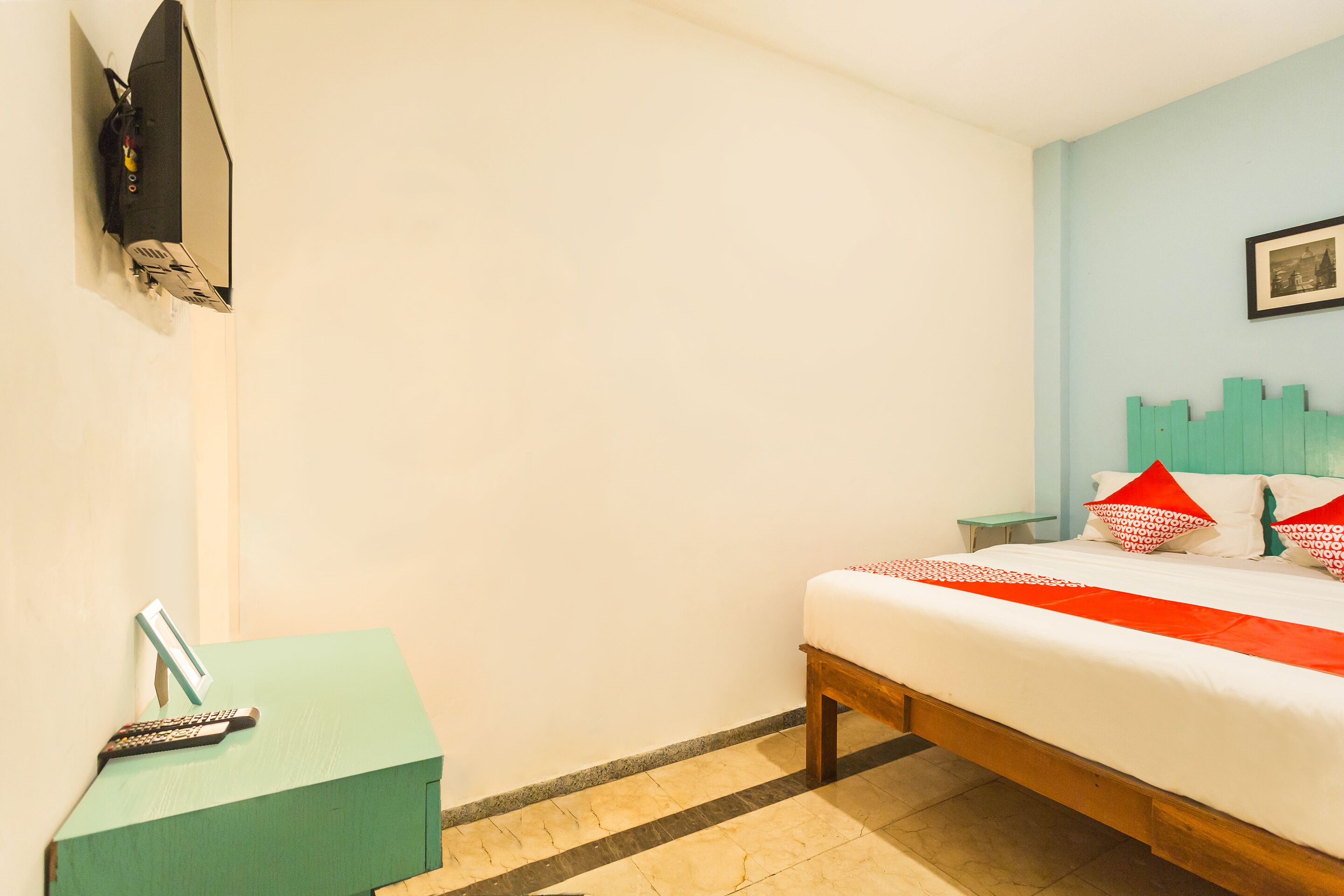 Quarto de hotel com cama, mesinha, televisão fixada na parede e um porta-retrato.