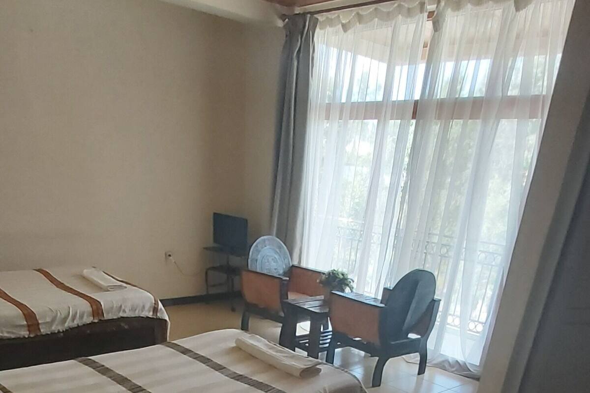 A hotel room with two beds, a desk with a chair, a small table, and a window with curtains.