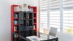 A modern room with a glass table, a red and black bookshelf, and a window with blinds. - Villa Ecume Des Jours (Les Terres Basses)