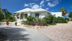 A white two-story house with a covered porch, surrounded by tropical plants and a sandy driveway. - Villa Ecume Des Jours (Les Terres Basses)