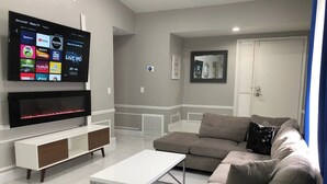 A modern living room with a flat-screen TV mounted on the wall, a white coffee table, a beige sofa, and a white cabinet with a TV stand. - 6183 Sandcrest · Walk TO Universal! Near Disney, Convention (Orlando)