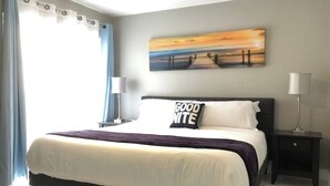 A neatly made bed with a white and purple bedspread, a pillow with "GOOD NITE" text, a painting of a sunset on the wall, and a nightstand with a lamp. - 6183 Sandcrest · Walk TO Universal! Near Disney, Convention (Orlando)