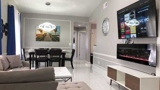 A modern living room with a flat-screen TV, a dining area, and a hallway. - 6183 Sandcrest · Walk TO Universal! Near Disney, Convention (Orlando)