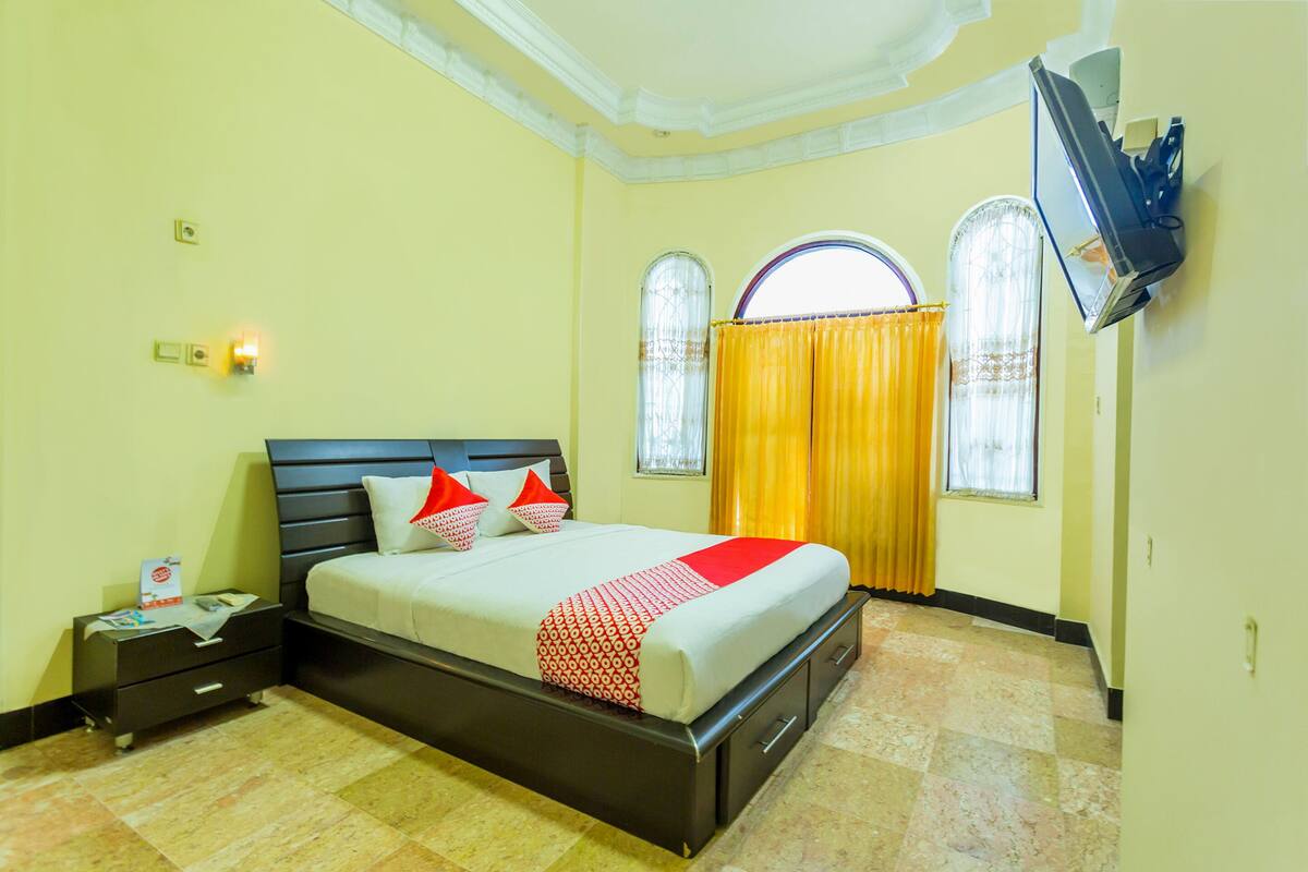 A hotel room with a bed, a nightstand, a TV mounted on the wall, and two windows with curtains.