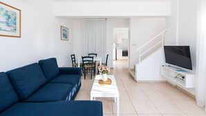 A modern living room with a blue sofa, a white coffee table with a vase, a flat-screen TV, and a staircase leading to a kitchen. - Fig Tree Z Holiday Villas DPS04-Two Bed Townhouse (Protaras)