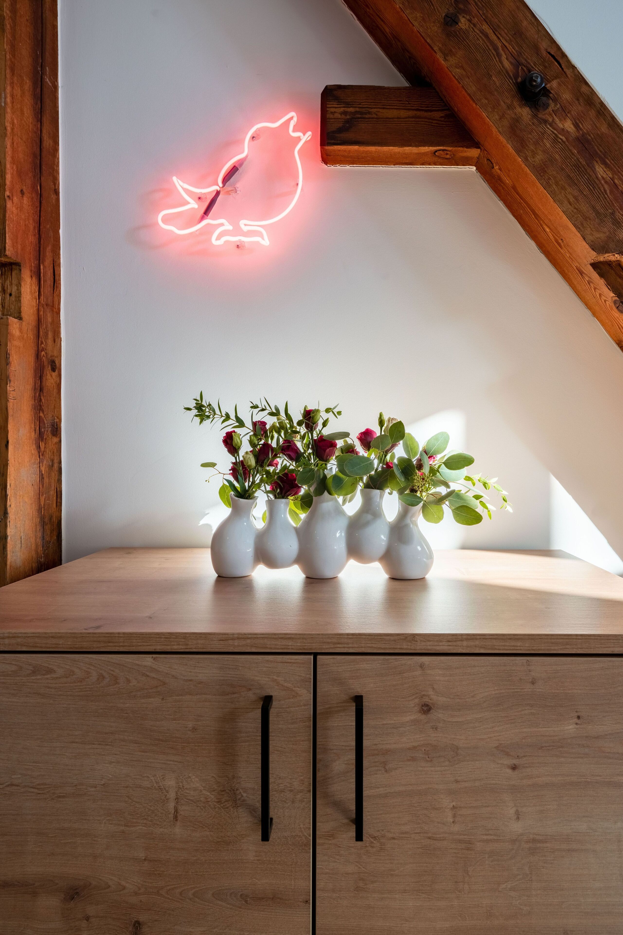 Un moble de fusta amb tres vasses blanques que contenen flors, un cartell de neon amb forma de parell i una estructura de bigues de fusta.