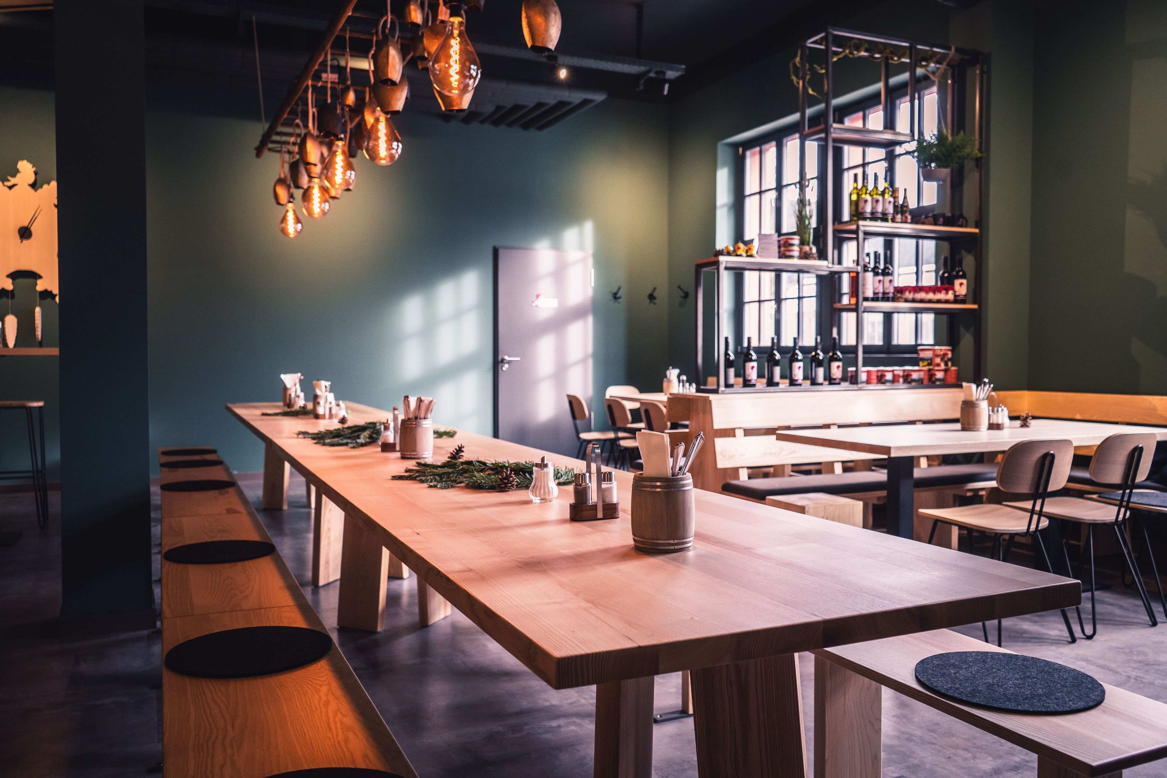 A modern restaurant with long wooden tables, hanging pendant lights, and a bar area with shelves of bottles.