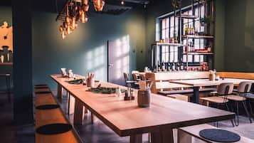 A modern restaurant with long wooden tables, hanging pendant lights, and a bar area with shelves of bottles.