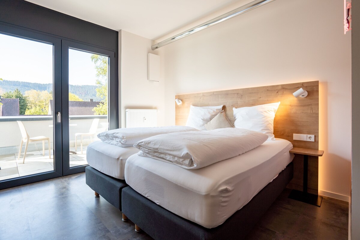 A double bed with a wooden headboard, a bedside table with a lamp, and a view of the outdoors through a sliding glass door.